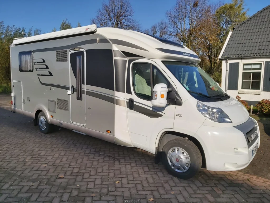 Ein silber-weißes Wohnmobil parkt auf einer gemauerten Auffahrt neben einem Haus mit dunklem Dach und weißen Wänden, umgeben von Bäumen - perfekt, wenn Sie Ihr gebrauchtes Wohnmobil verkaufen oder ein zuverlässiges Wohnmobil ankaufen wollen.