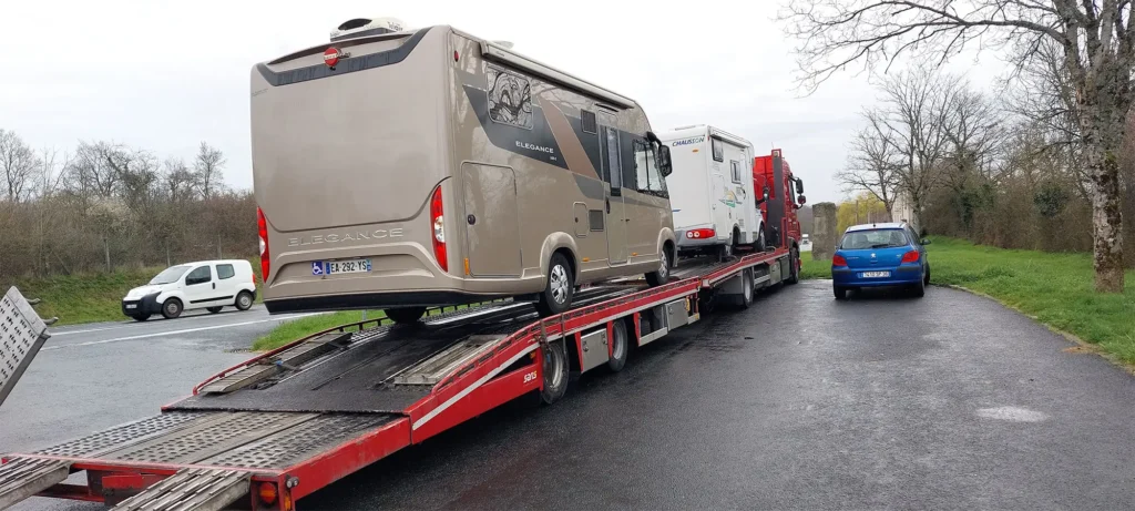 Ein Autotransporter-Anhänger mit zwei Wohnmobilen und einem Kleinwagen parkt am Straßenrand neben einem blauen Auto und Bäumen - ideal für alle, die wohnmobil verkaufen oder wohnmobil ankaufen wollen.