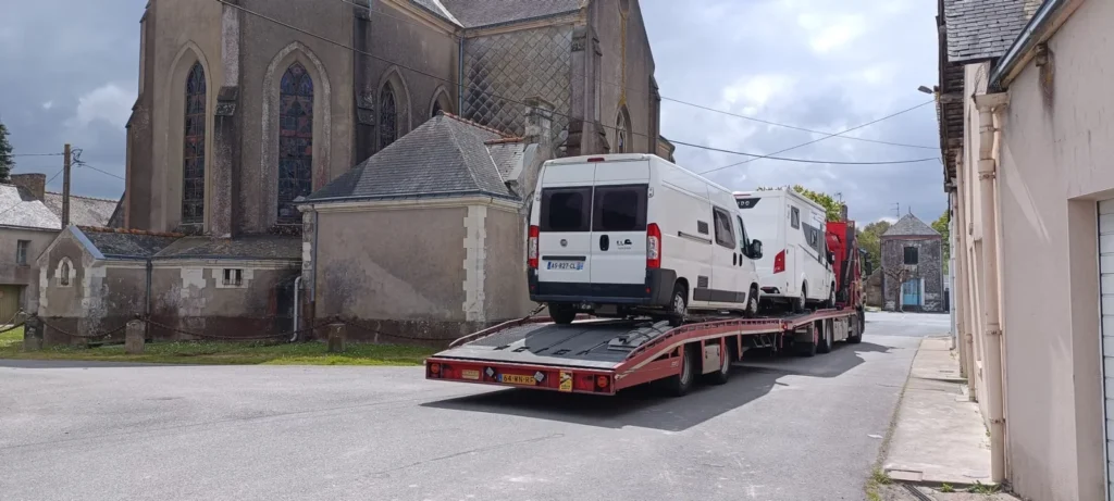 Ein Pritschenwagen transportiert zwei weiße Lieferwagen auf einer Straße in der Nähe alter Steinbauten und einer Kirche mit Buntglasfenstern - ein idealer Ort, um wohnmobil zu verkaufen oder Tipps zum Verkauf des Wohnmobils zu entdecken.