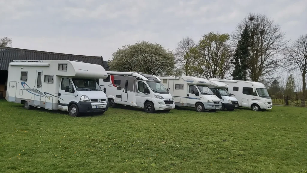 Fünf Wohnmobile stehen in einer Reihe auf einer Wiese in der Nähe einiger Bäume und eines Gebäudes unter einem wolkenverhangenen Himmel - die perfekte Inspiration, wenn Sie an wohnmobil verkaufen oder wohnmobil ankaufen denken.