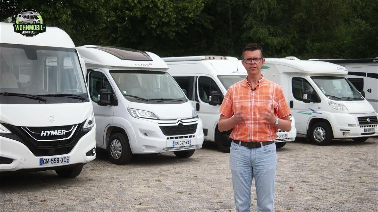 Eine Person in einem orangefarbenen Karohemd steht vor einer Reihe geparkter weißer Wohnmobile auf einem gepflasterten Platz, im Hintergrund sind Bäume zu sehen.
